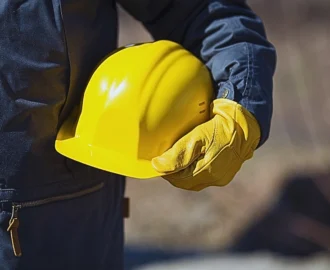 A person wearing a dark jacket and yellow protective gloves holding a bright yellow construction helmet. Delays in Work Permit Renewals