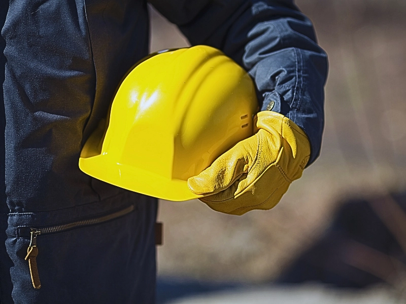 A person wearing a dark jacket and yellow protective gloves holding a bright yellow construction helmet. Delays in Work Permit Renewals