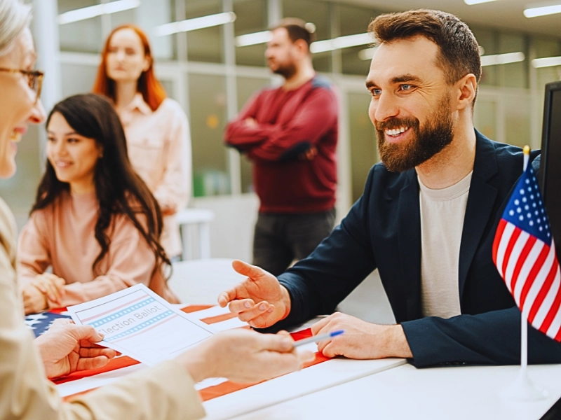 a man with a beard sitting at a registration table, engaging in conversation. Immigration Parole Programs