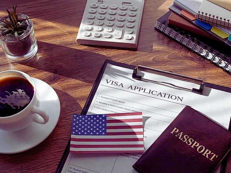 a visa application form on a clipboard, accompanied by a small American flag card and a passport. H-1B and L-1 Visas